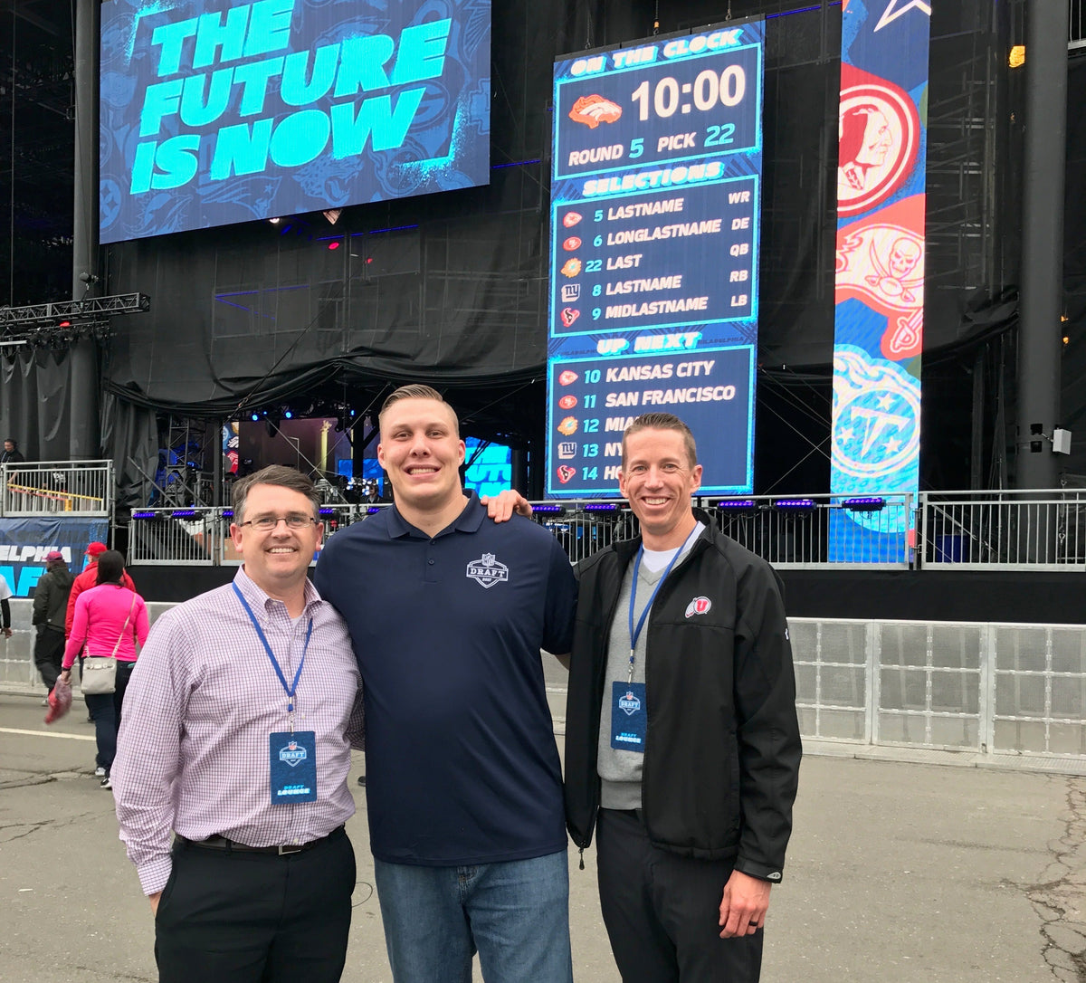 Three people standing in front of a large screen displaying 'The Future is Now' at an event.